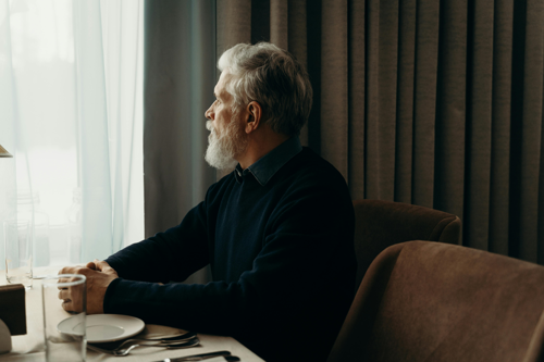 A photograph of an older looking man with a beard looking out of his window